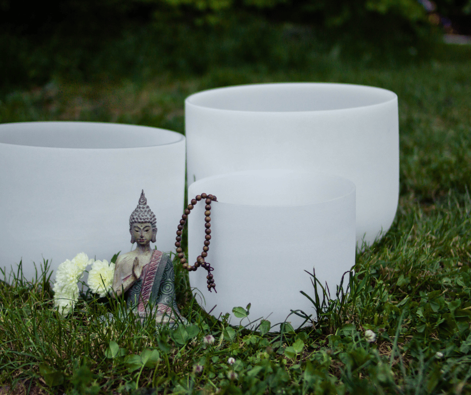 Three crystal singing bowls on grass with a buddha statue and wooden beads